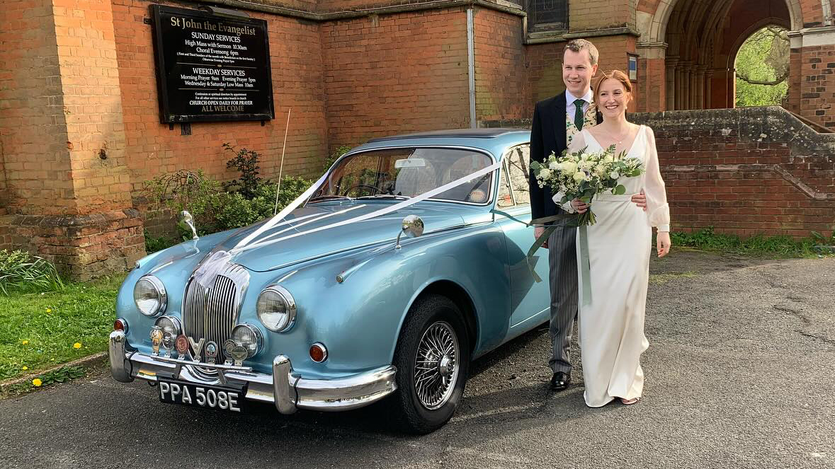 Silver Blue Classic Daimler 250 V8 decorated with white ribbons. Bride holding a bouquet of flowers and Groom are standing by the vehicle