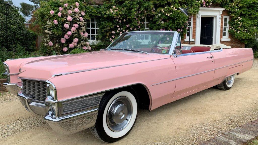 Pink Cadillac convertible with roof down and white wall tires on front of a wedding venue with pink flowers in the background.