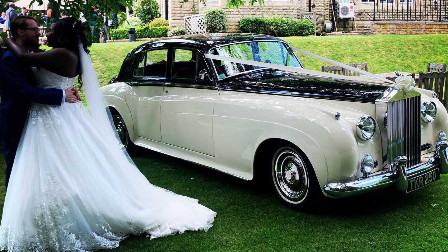 Black & Ivory Classic Rolls-Royce Silver Cloud in the Garden of the venue with Bride and Groom stnading by the vehicle kissing each others.