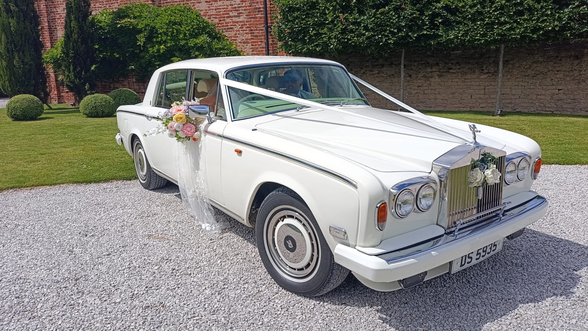 classic White Rolls-Royver silver Shadow decorated with white ribbons and flowers. Bride and Groom can be seen inside the vehicle.