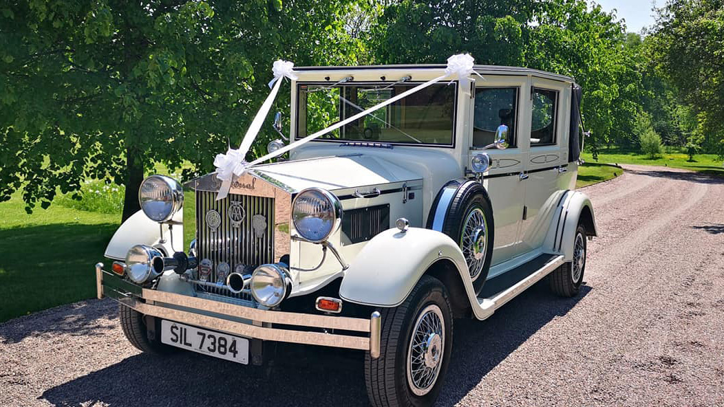 Vintage 1930s style Imperial convertible with roof close decorated with traditional white wedding ribbons and bows across its bonnet. Vehicle is parked on the drive of a local Barn
