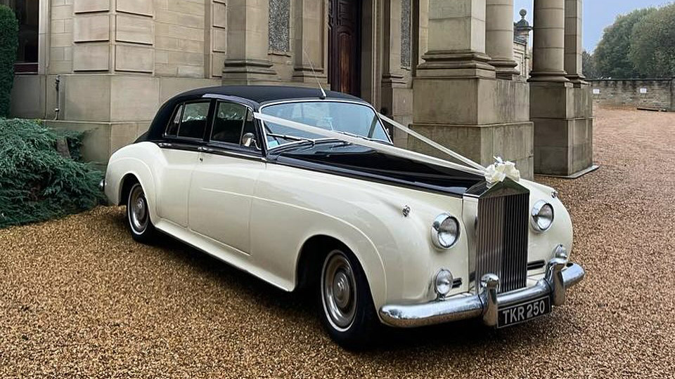 classic White Rolls-Royce Silver Cloud with black roof and bonnet decorated with white ribbons and bow parked in front of wedding venue.