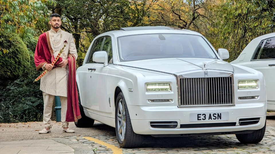 A modern white Rolls-Royce Phantom with headlights on and Groom stands by the vehicle with his traditional Asian outfit and sword.