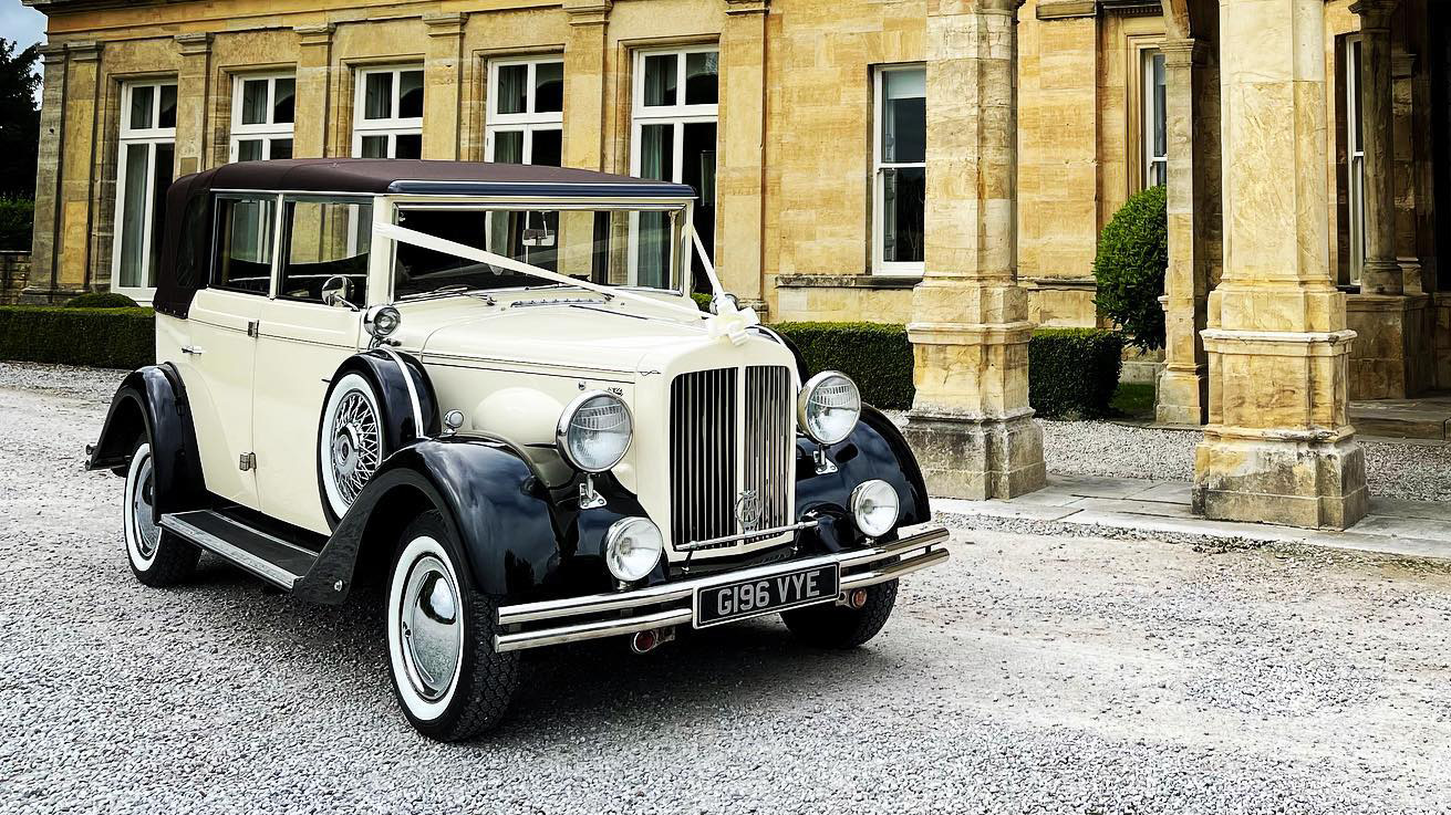 1930s vintage regent in Ivory and Black parked in front of a wedding venue. Vehicle is dressed with Ivory ribbons and bow and the convertible part of the roof is up.