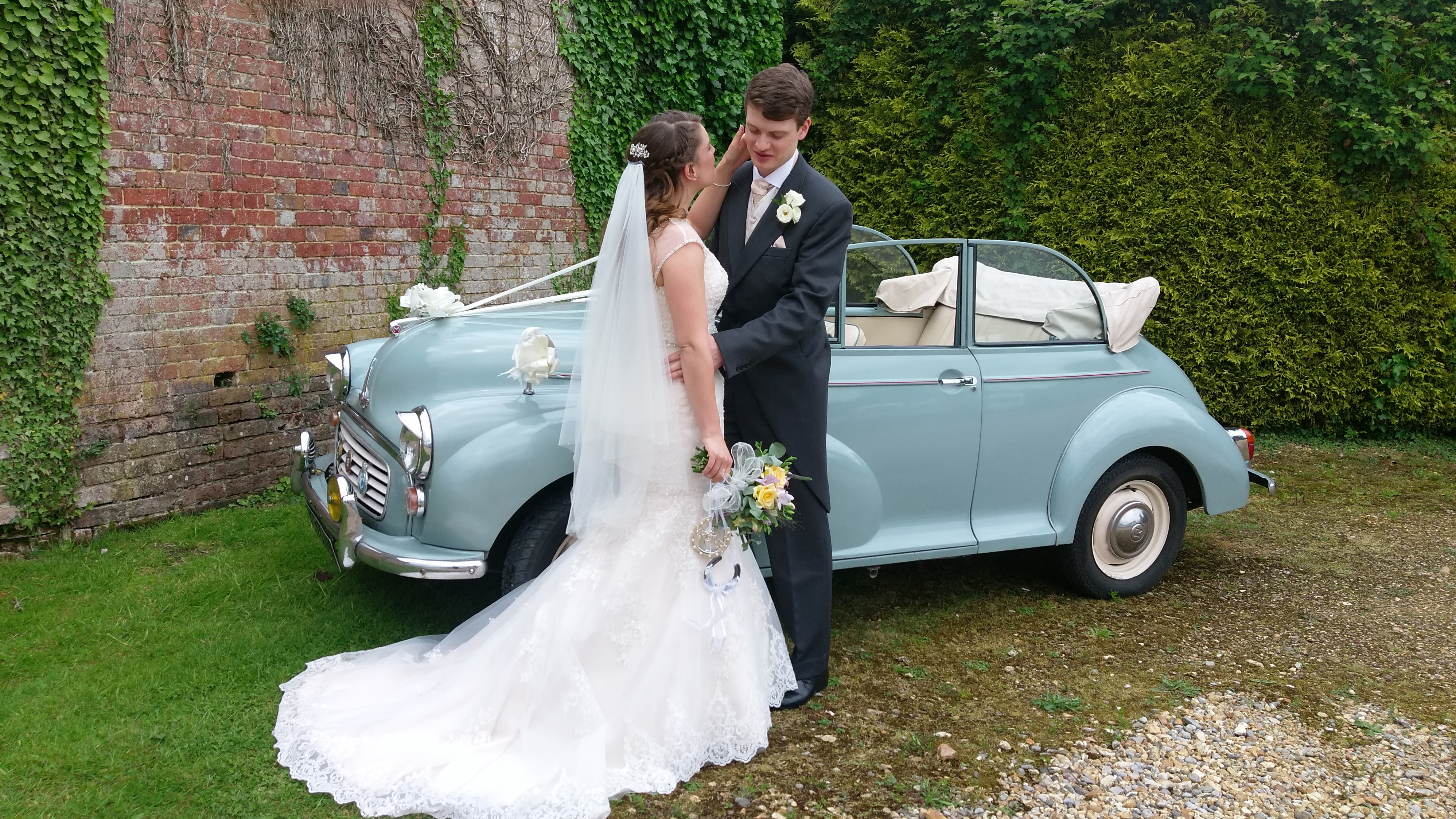 Lymington-based Classic Morris Minor with ribbons and bow in light blue with its roof open showing a light cream interior. Bride and Groom are in front of the vehicle posing for their wedding photographer.