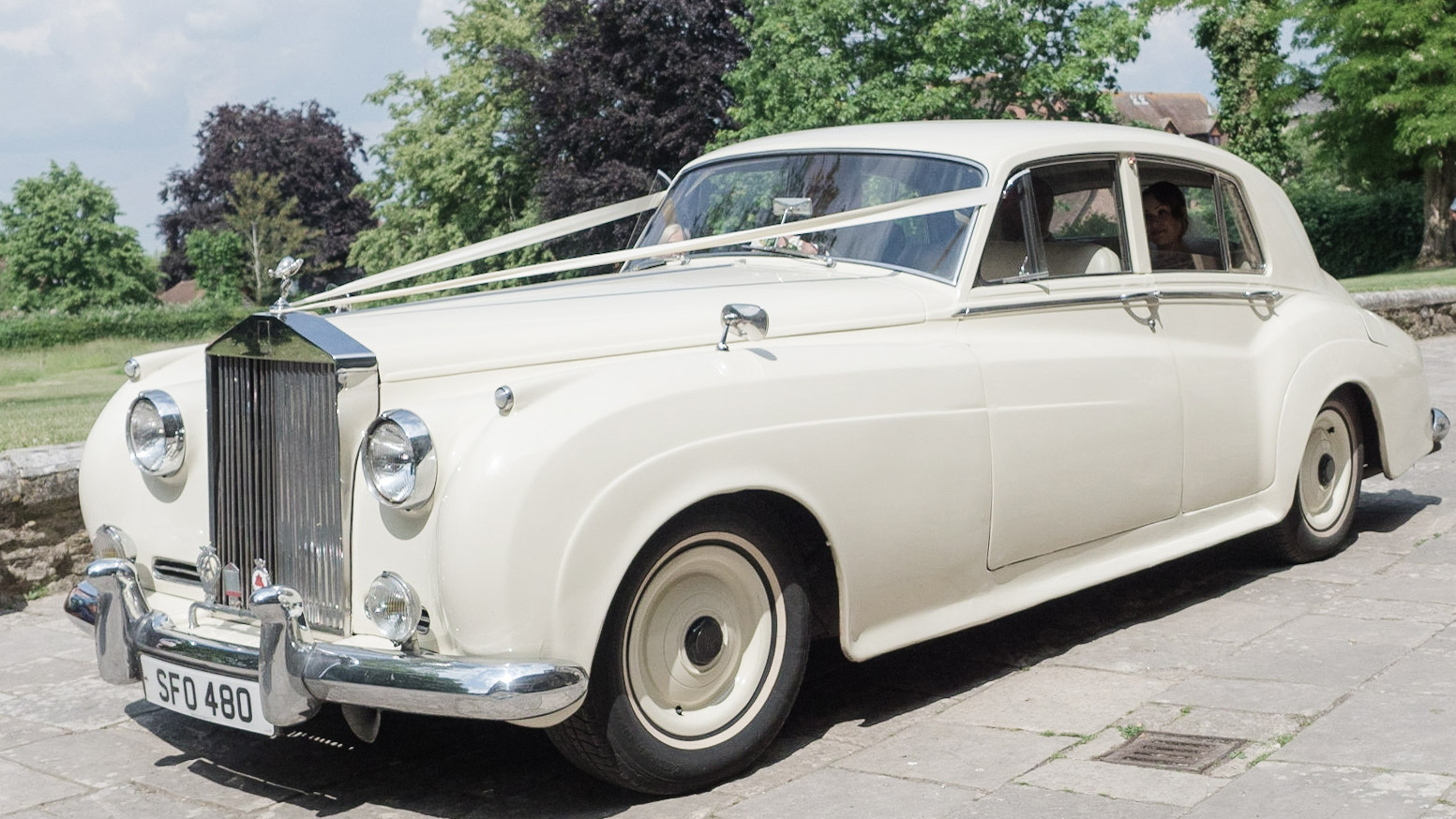 Classic Old English White Rolls-Royce Silver Cloud decorated with Ivory ribbons. Bride can be seen at the rear of the vehicle and chauffeur at the front