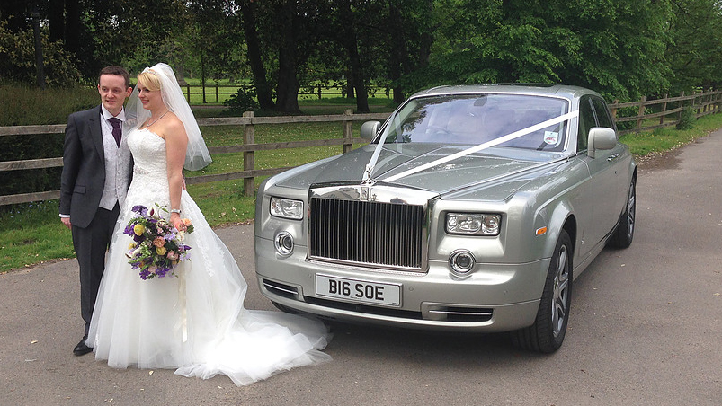 A Silver Rolls-Royce Phantom series 2 with white ribbons across the bonnet. Bride and Groom are standing in front of the vehicle for photos. Bride is holding a large bouquet made o
