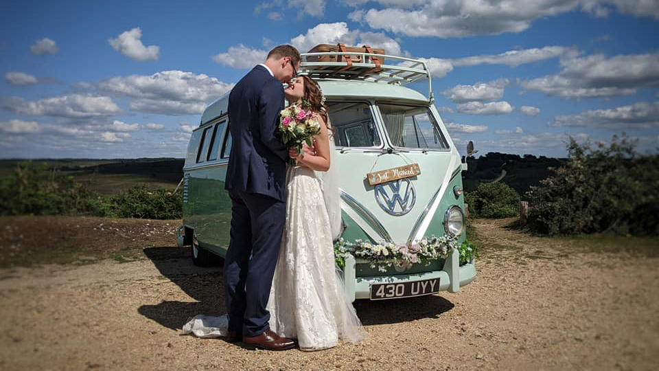 Retro Volkswagen Campervan Splitscreen teo tone White and Pastel Green decorated with white ribbons, green foliage on front bumper and a picnic basket on top of its roof. Bride and Groom are standing in front of the vehicle kissing.