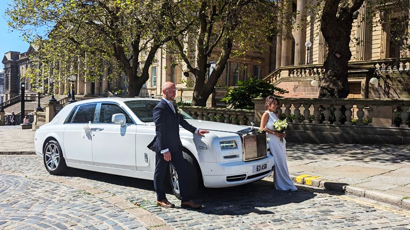 A modern white Rolls-Royce Phantom parked in the street of Wetherby with Bride and Groom posing for their wedding photographer in front of the vehicle.
