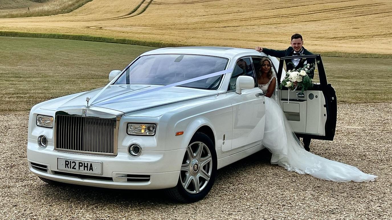 White Rolls-Royce Phantom with traditional White ribbons with Bride and Groom. Bride is seating in the rear seat with her legs outside the vehicle white the groom is holding the backward opening door for his Bride.