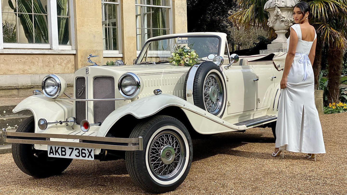 Ivory vintage Beauford with soft top down and spare wheel mounted on side skirt. Bride wearing a white dress standing by the vehicle at a local wedding venue inLowestoft.