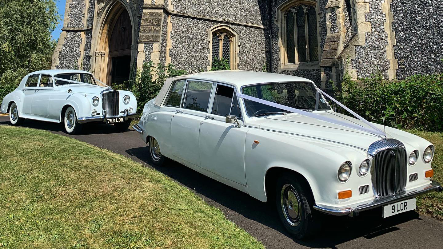 A classic Daimler Limousine in white followed by a Rolls-Royce Silver Cloud both decorated with matchign pair of white ribbons parked in front of a church inWoodbridge.