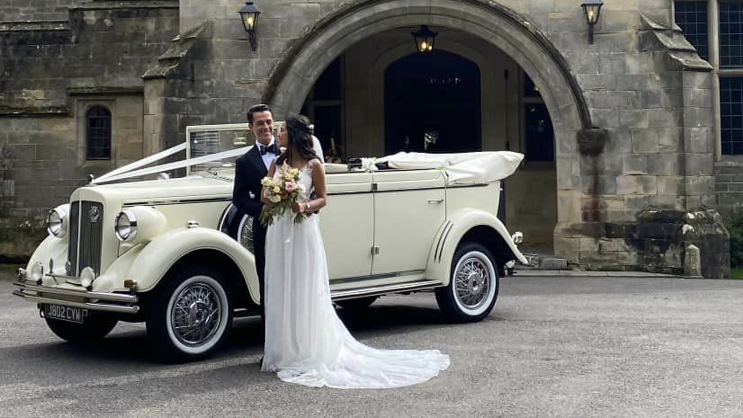 Full Convertible Regent in ivory decorated with traditional white wedding ribbons. Bride and Groom are standing next to the vehicle holding and smiling at each others. Vehicle is p