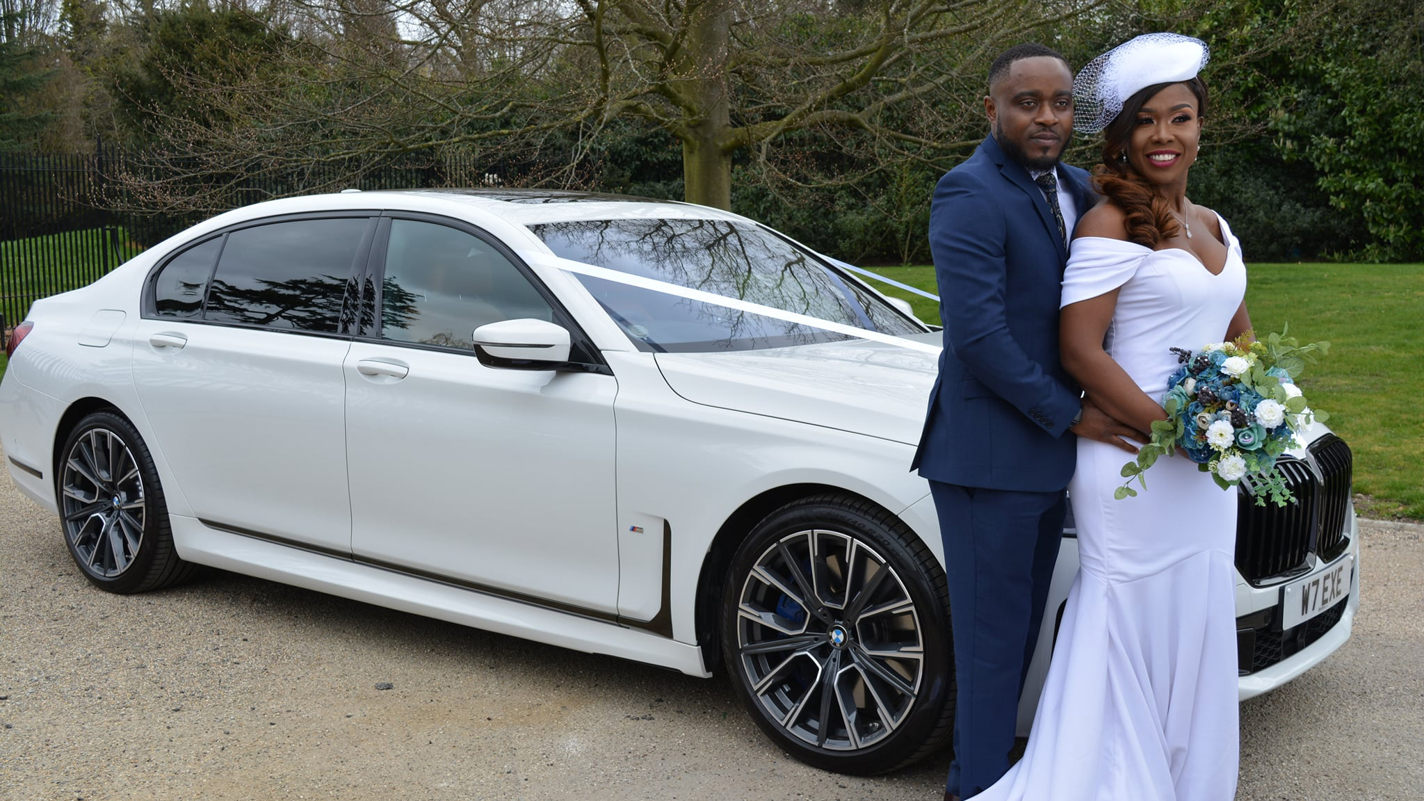 White BMW decorated with white ribbons. Bride and Groom are standing by the vehicle smiling and holding each others.