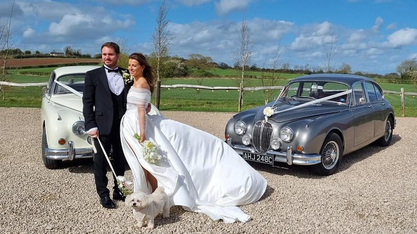 A classic silver Jaguar Mk2 and an Ivory Daimler 250 V8 dressed with matching White ribbons accros the front bonnet. Bride and Groom are standing in the middle of the two vehicles.A classic silver Jaguar Mk2 and an Ivory Daimler 250 V8 dressed with matching White ribbons accros the front bonnet. Bride and Groom are standing in the middle of the two vehicles.