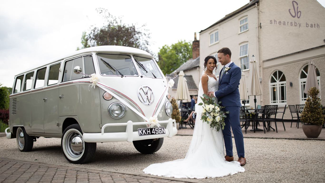 Classic VW Campervan in silver and white dressed with white ribbons and bows. Bride and Groom are holding each others in front of the vehicle. Shearsby Bath wedding venue in Leicestershire can be seen in the background.