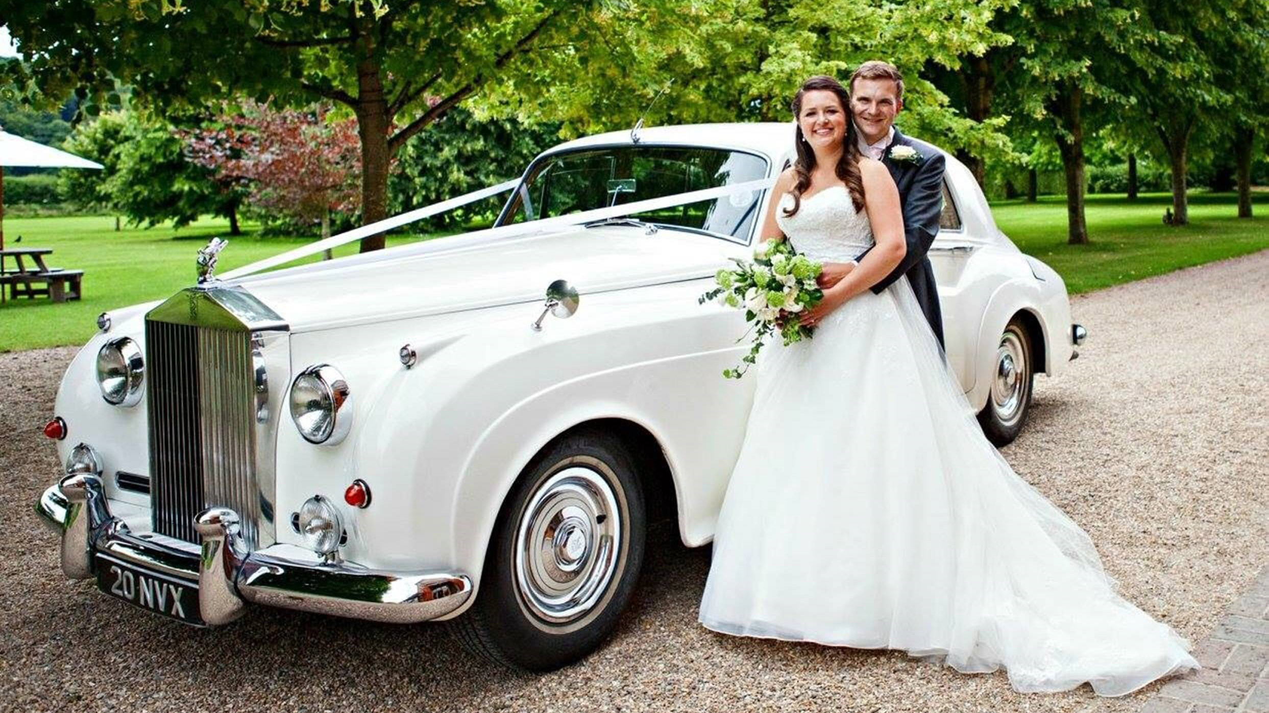 White Rolls-Royce Silver cloud decorated with ivory ribbons parked in the entrance of a wedding venue in Barton-le-Clay. Bride and Groom are standing by the vehicle. Groom is holding his Bride in his arms while she holds her bridal bouquet.