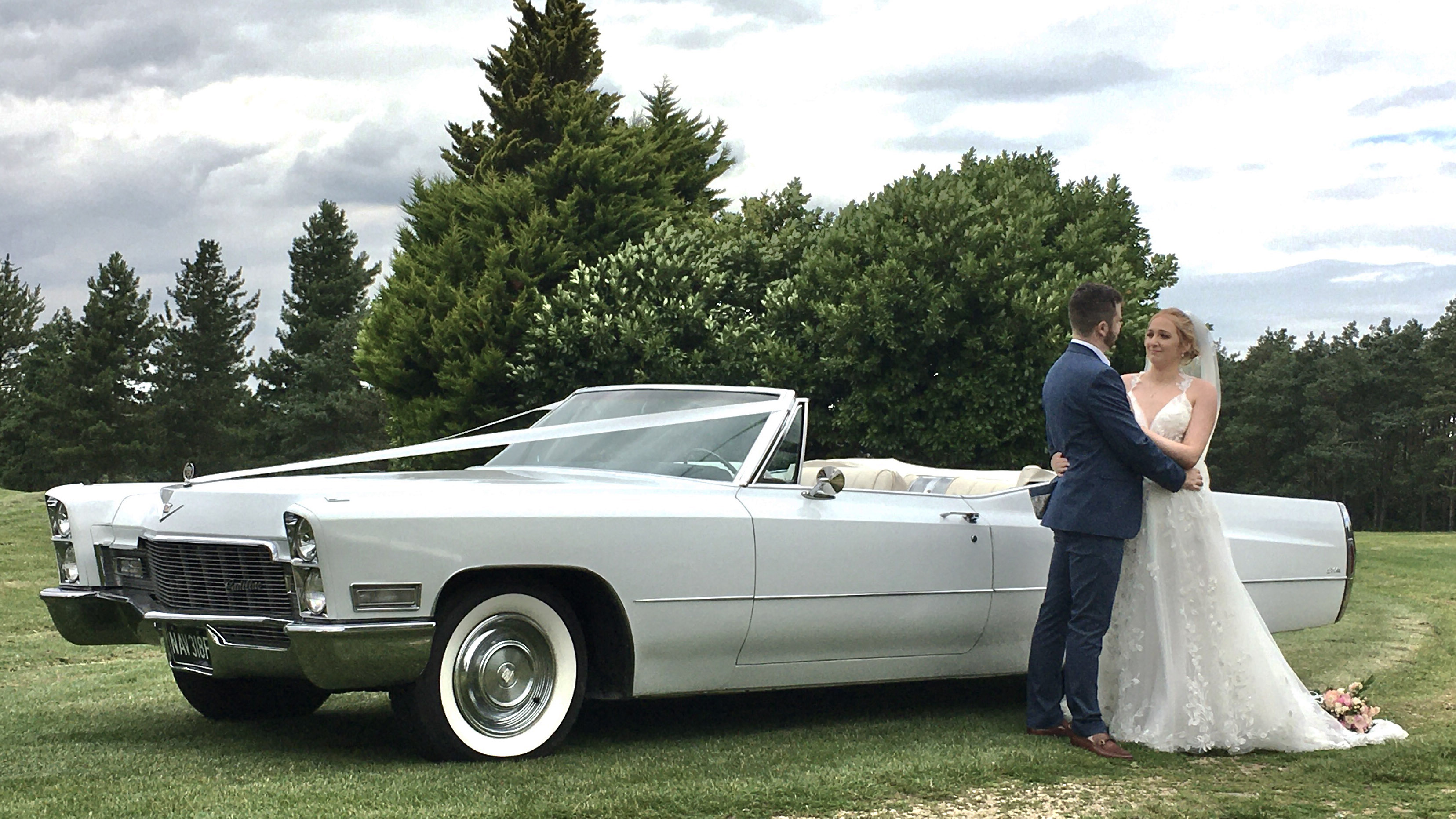 White American Classic Cadillac in white with roof down, decorated with white ribbons. Bride and Groom re standing by the vehicle holding and looking at each others.