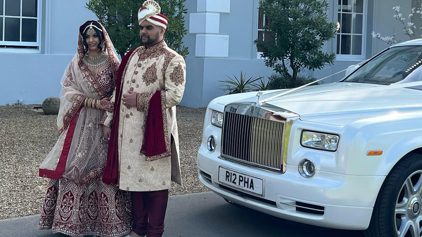 Asian Bride and Groom wearing traditional Burgundy and Gold ceremonial outfits in front of a wedding venue in Sheffield with a white Rolls-Royce Phantom.
