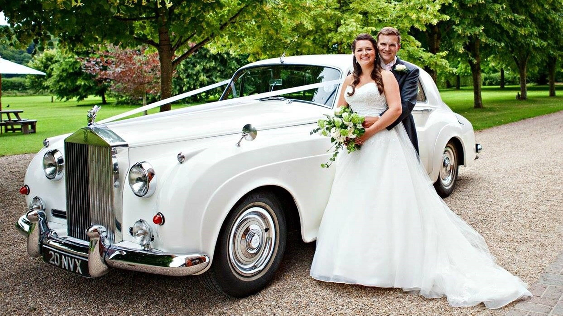 White Rolls-Royce Silver cloud decorated with ivory ribbons parked in the entrance of a wedding venue in Solihull. Bride and Groom are standing by the vehicle. Groom is holding his Bride in his arms while she holds her bridal bouquet.