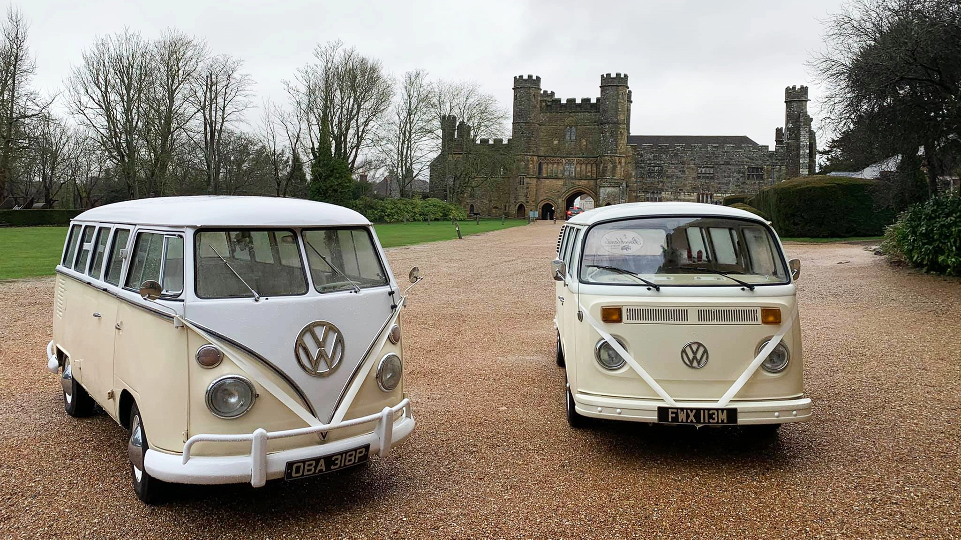 Two classic Stourbridge-based VW Campervans in Cream and White decorated with matching white ribbons at the end of a drive way with the wedding venue in the background.