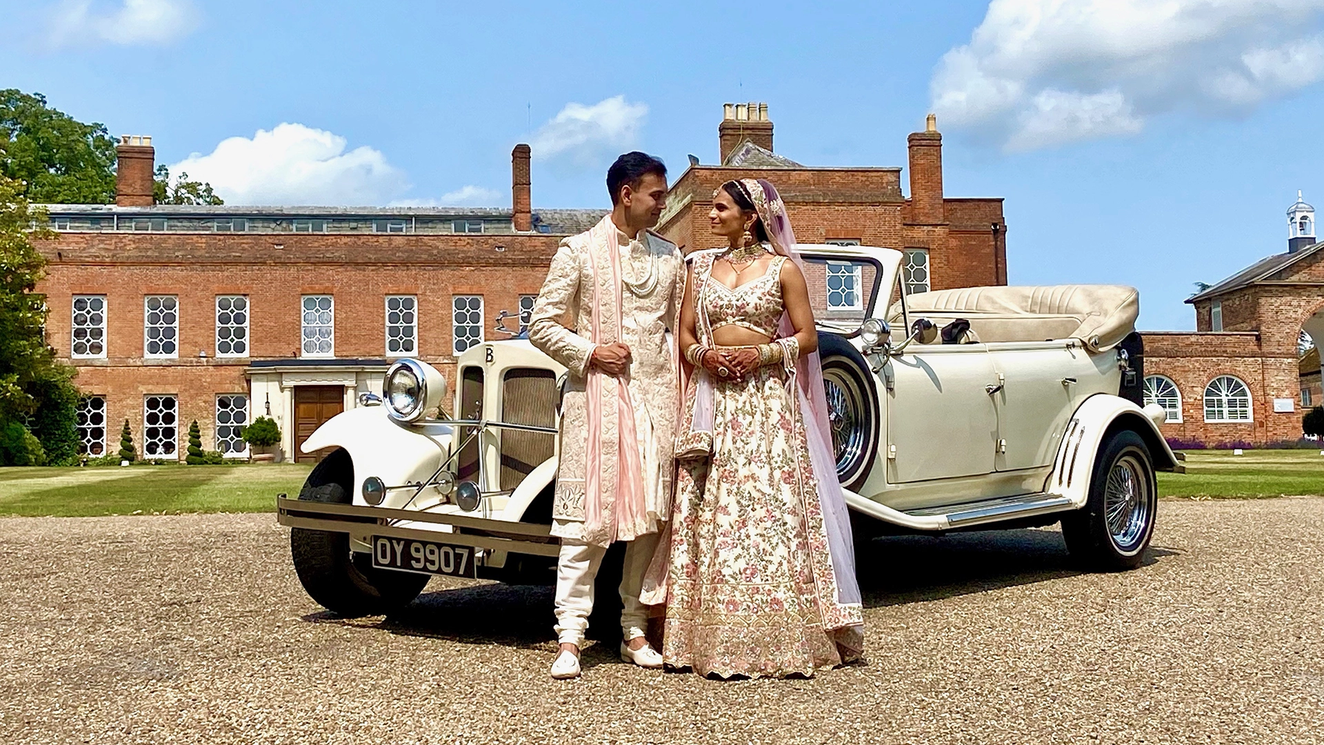 1930's vintage style Beauford in ivory with roof down. Asian Bride and Groom in their traditional outfit standing in front of the vehicle. Local Cumbria venue in the background.