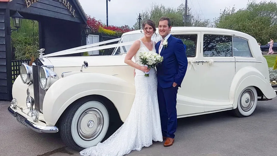 Rolls-Royce Silver Wraith decorated with Ivory ribbons. Bride and Groom are standing in front of the vehicle posing for their wedding photographer