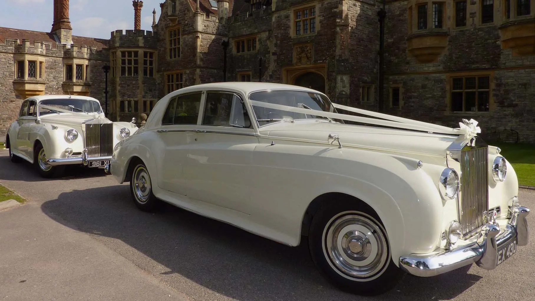 Ivory classic 7-seater Austin Princess Limousine dressed with a traditional V-shape ivory ribbons across its front bonnet. Vehicle is standing in the middle of a path in a Colchest