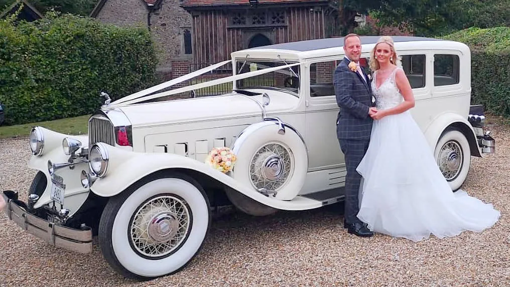 a Vintage Pierce Arrow Limousine decorated with ivory ribbons, Large banded white walltires with spokes wheels. Bride and Groom are standing in front of the vehicle holding each others.