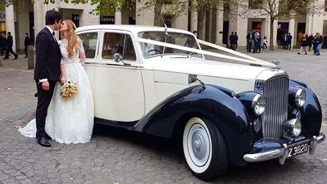 Classic Bentley Mk6 decorated with white ribbons and Bride and Groom kissing next to the vehicle