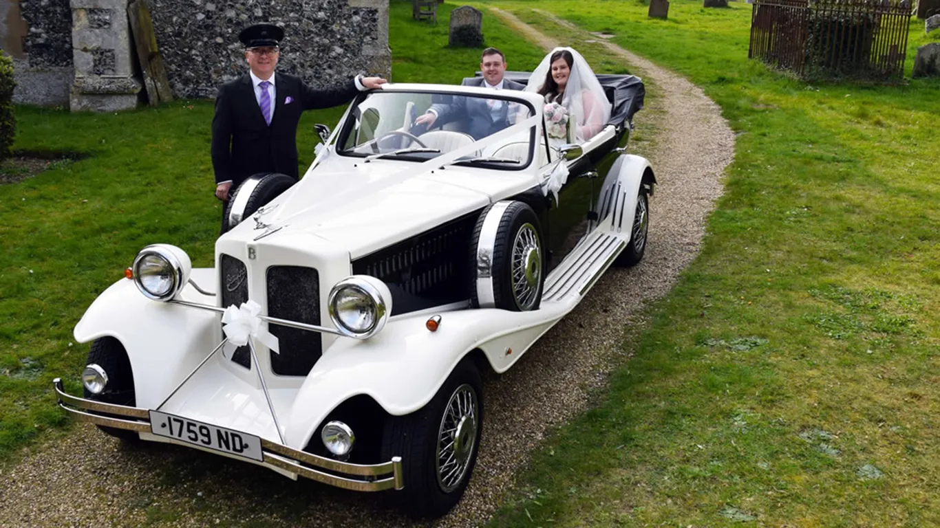Black & White Beauford with Chauffeur Standing next to the car with his hat, dark grey suit and Purple Tie and the Bride and Groom seating in the vehicle