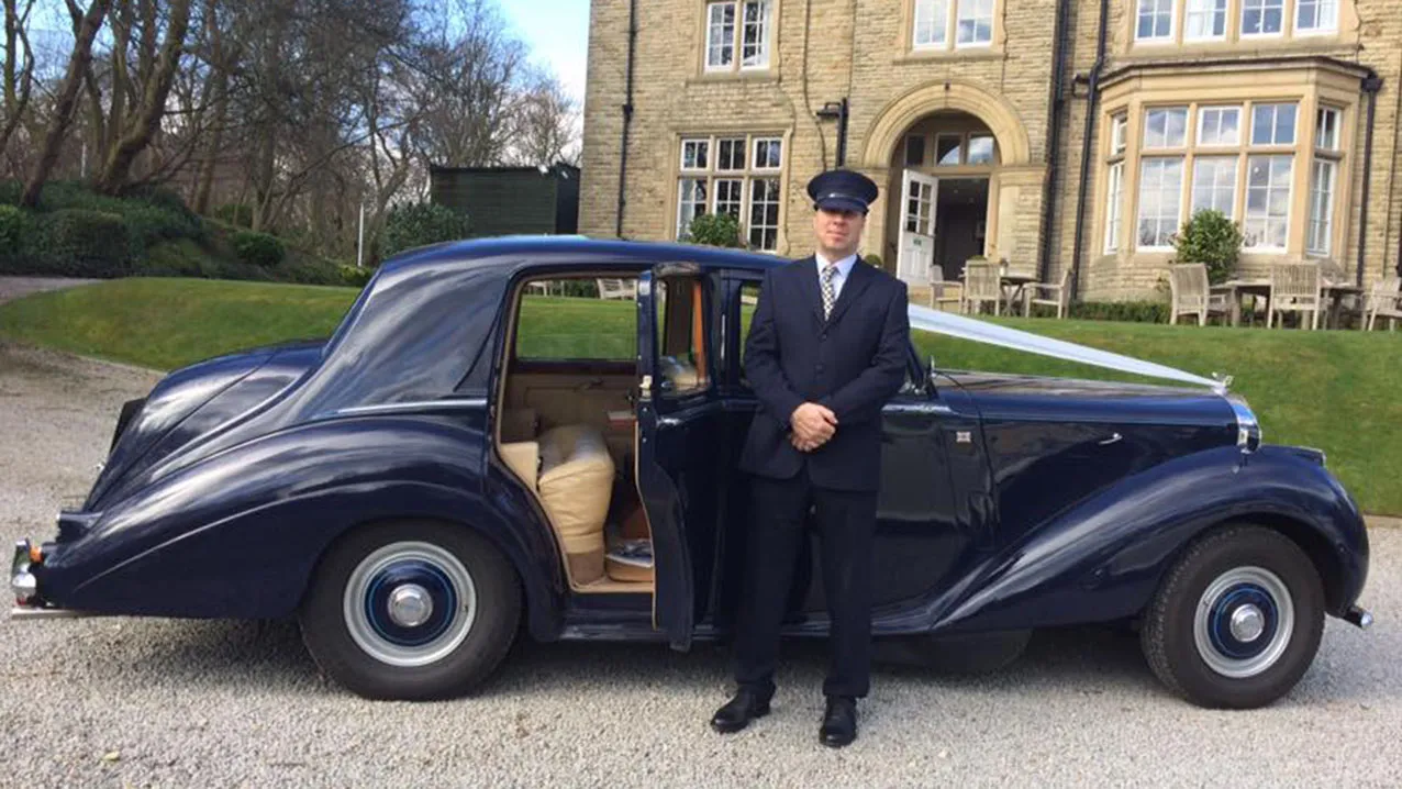 Fully Uniformed Wedding Chauffeur with Grey Wedding Suit and Chauffeur's Hat standing in front of a Dark Blue Classic Bentley
