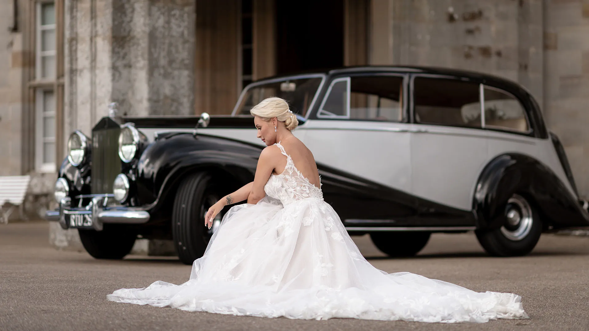 Bride dearing a large white wedding dress seating in front of a classic Rolls-Royce in two-tone Black and Silver