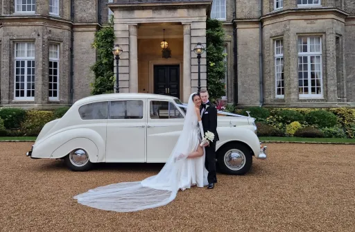 Classic Austin Princess in white decorated with white ribbons in front of wedding venue in Cambridgesire. Bride and Groom standing in front of the vehicle holding hands and posing for photo