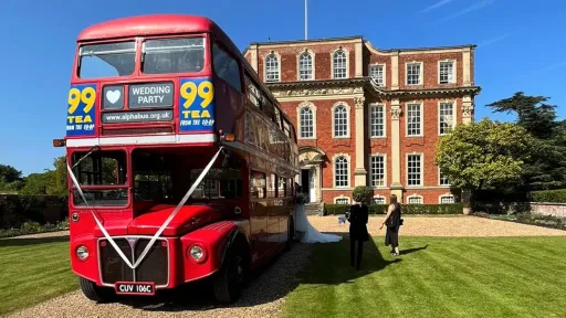 Vintage Red Routemaster bus dropping guests off at a Kent wedding venue seen in the background.  Bus is decorated with White ribbons and displays vintage advertising on its side.