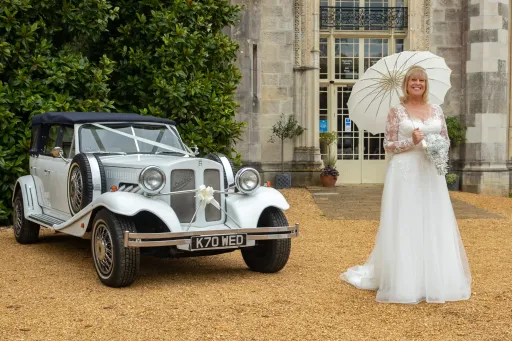 Vintage Rolls-Royce with White Ribbons and Bride standing in front of the vehicle. Bride is wearing a white dress and holding a white umbrella