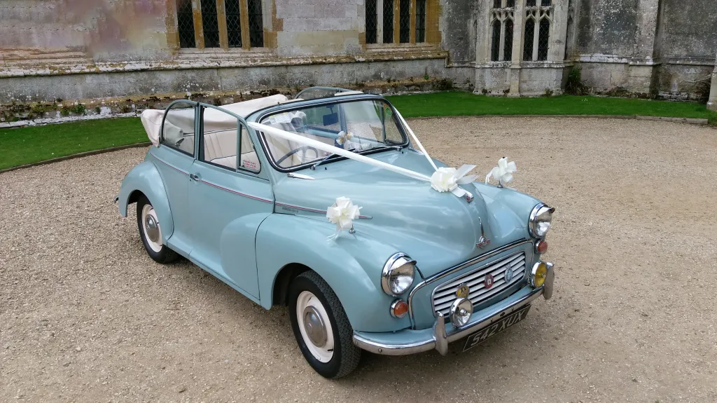 Pale Blue Classic Morris Minor Convertible with roof open showing the White leather interior and dressed with White Wedding Ribbons and Bows
