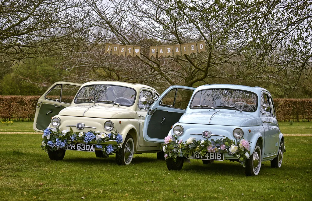 Two Classic Fiat 500 in Blue and ivory with Flower Decoration on front bumper. Both Vehicles have a door open. "Just Married" banner above the vehicles