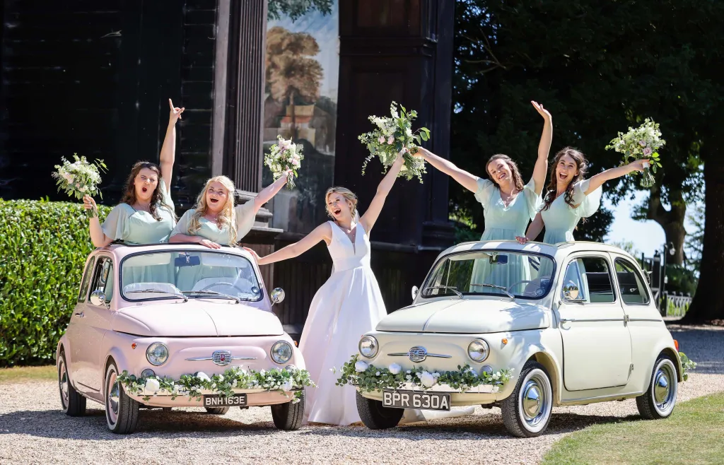 Two Classic Fiat 500 Wedding Cars. A pink Fiat and the other in ivory. Bridesmaids are standing in the vehicles and bride is in the middle of both vehicles all waving with flowers in their hands