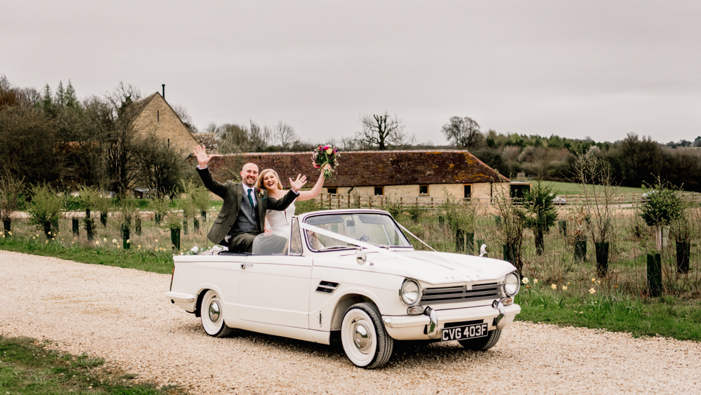 White Convertible Trimph with bride and groom seating in rear seat and waving