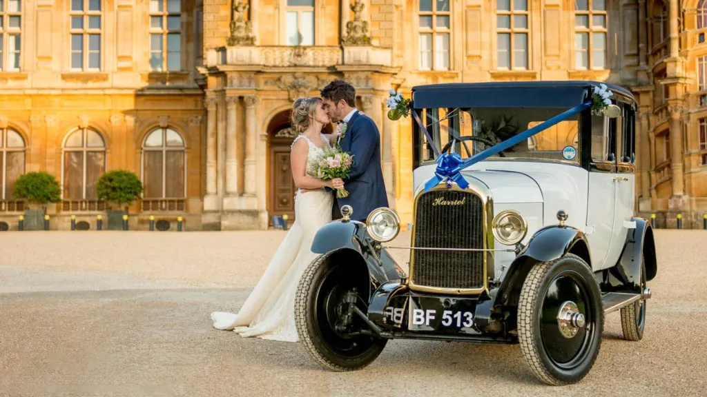 Vintage Citroen dressed with Royale Blue Ribbons in front of wedding venue with Bride and Groom standing next to the vehicle kissing