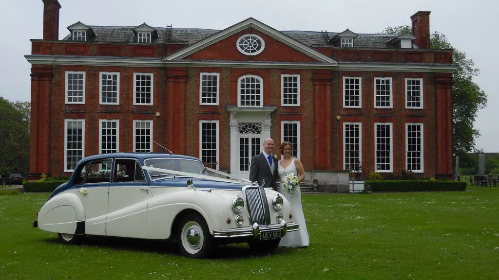 Classic Armstrong Siddeley in Ivory & Blue in front of a wedding venue. Bride and Groom are standing next to the vehicle posing for their wedding photographer.