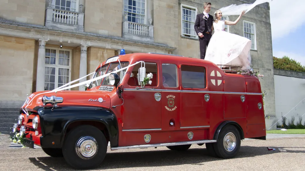 Side view of a Red Fire Engine decorated with wedding ribbons andf flowers. Bride and Groom standing on the vehicle