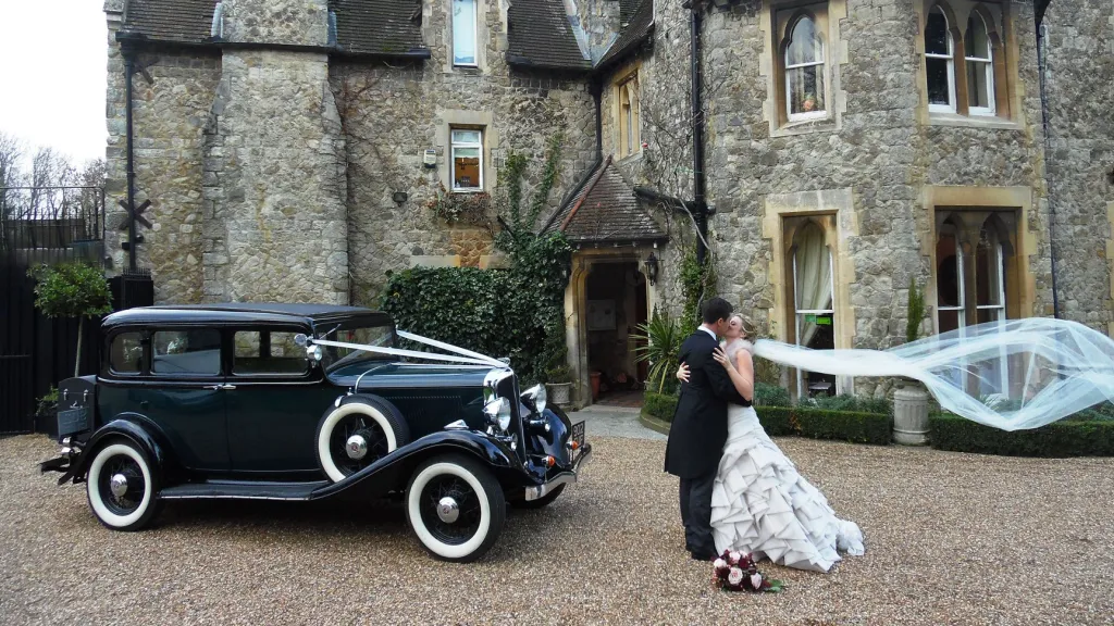Dark Green Vintage Studebaker car in front of wedding venue with Briode and Groom kissing next to the vehicle