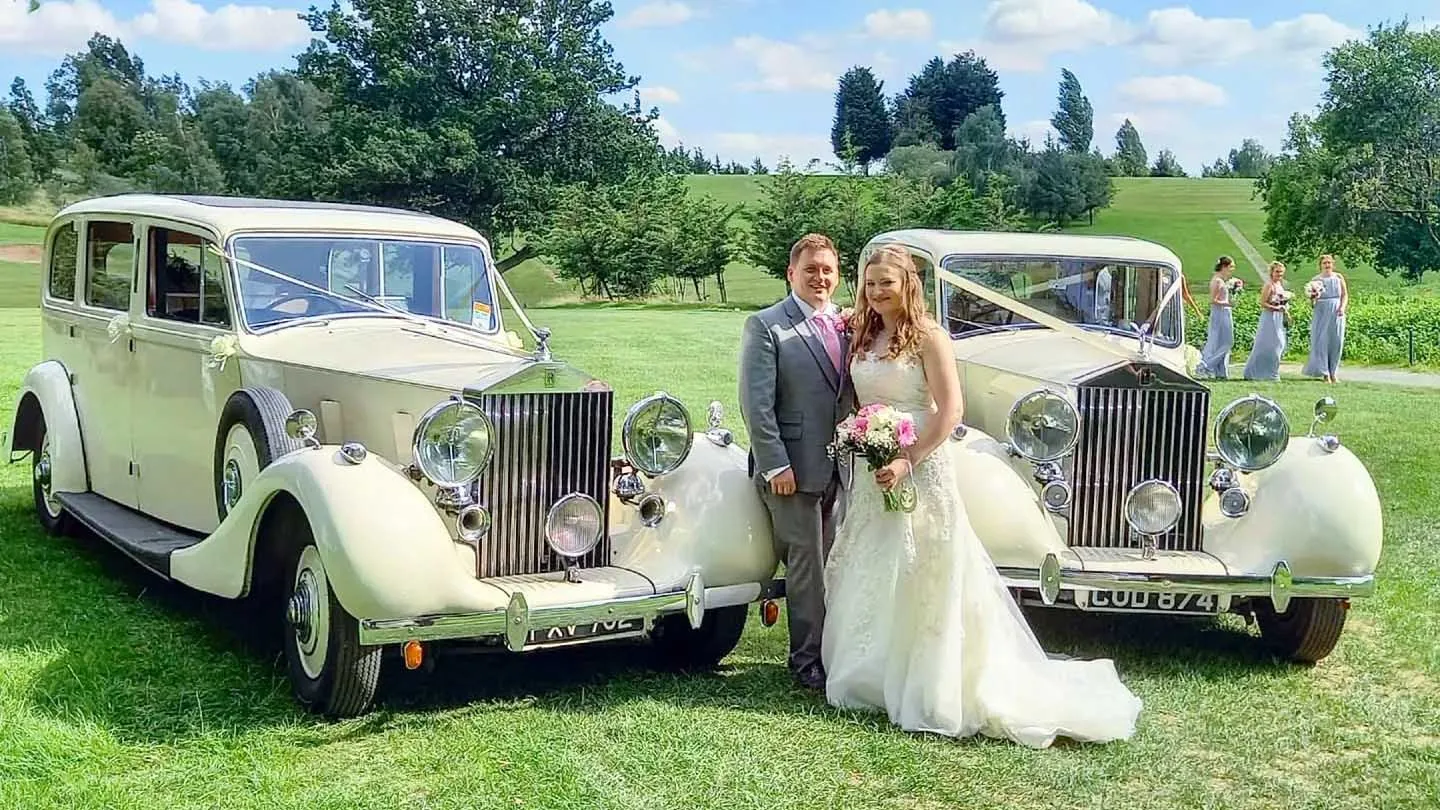 Matching Vintage Rolls-Royce Limousine with Bride and Groom standing in the middle of the two vehicles. Bridesmaids walking in their blue dress in the background