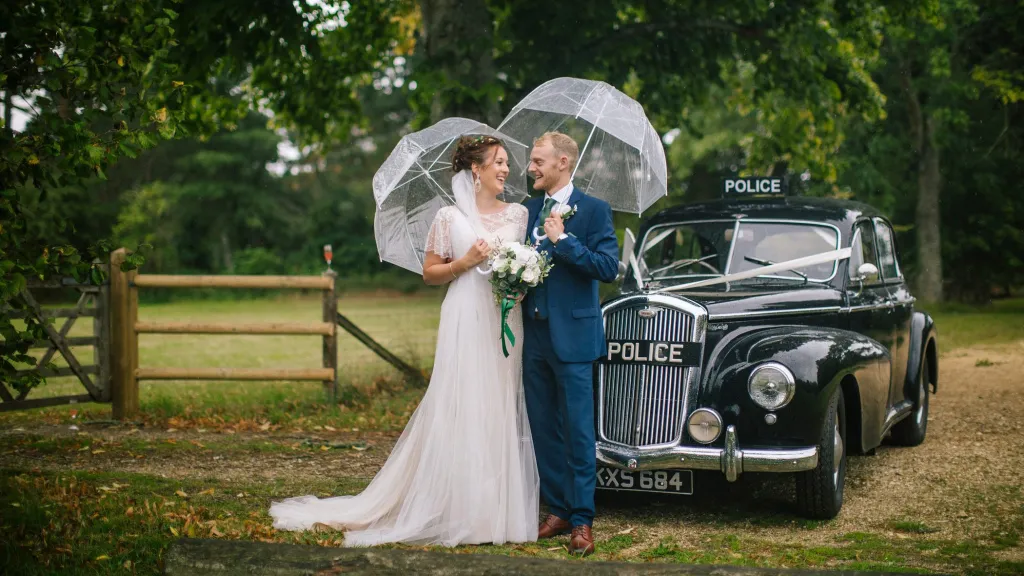 Black Classic wolseley Police Car with bride and groom standing in front of the vehicle with umbrellas