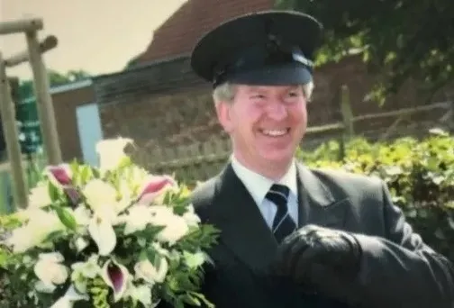 A smiling wedding chauffeur dressed with Dark Grey Suit and Chauffeur's Hat holding some flowers.