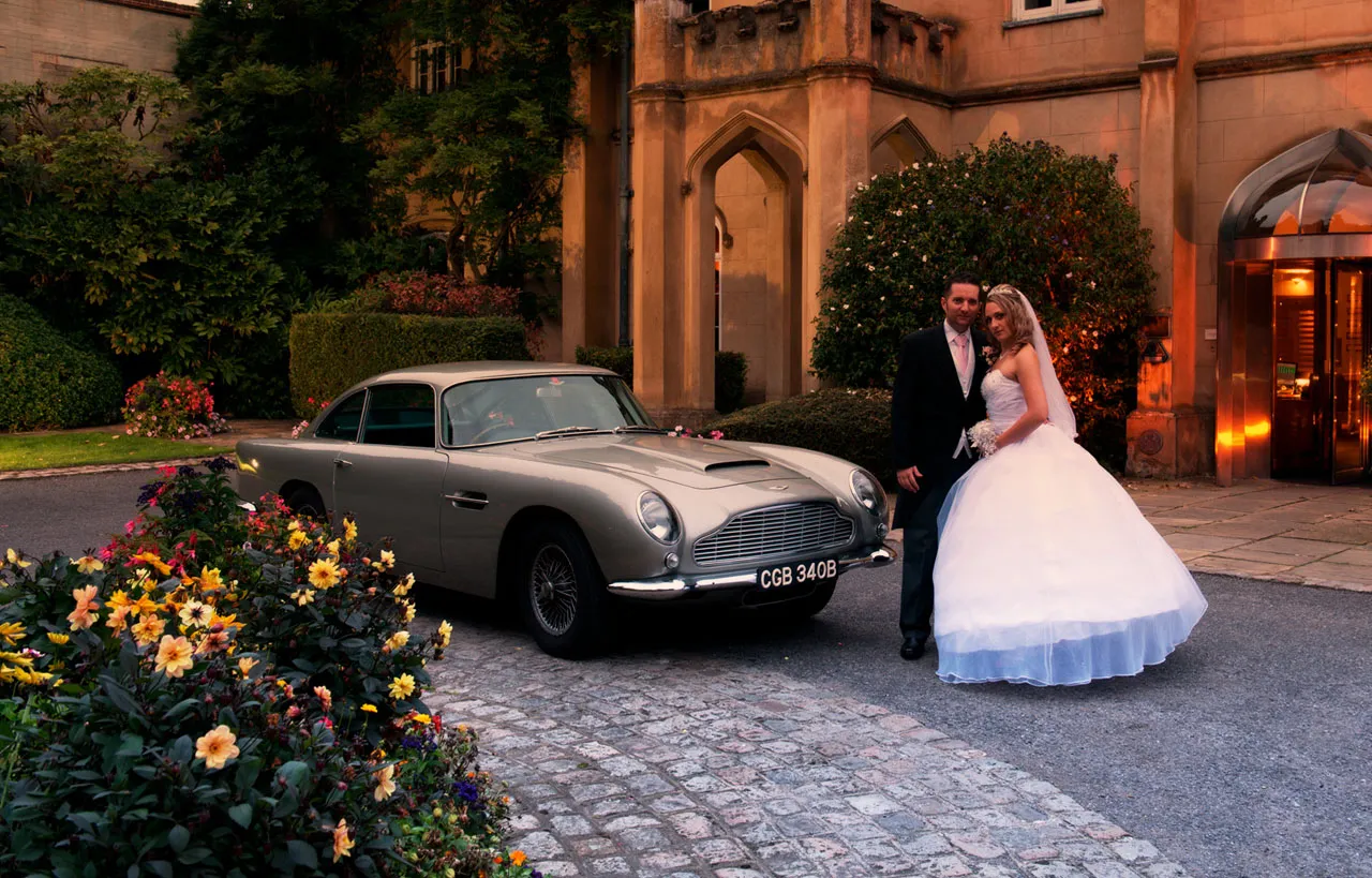 Classic Aston Martin DB5 in front of wedding venue at Dusk with Bride and Groom posing in front of the vehicle