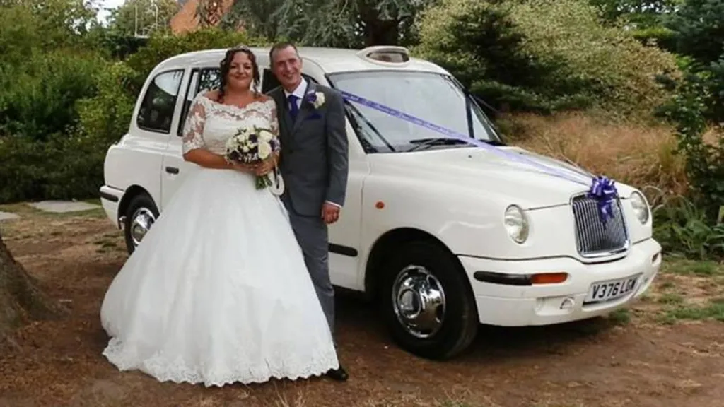 White Taxi Cab decorated with Purple Ribbons and a bow on its front grill. Bride and Groom are standing next to the vehicle posing for their wedding photographer. Bride is holding a bouquet of flowers in her hands