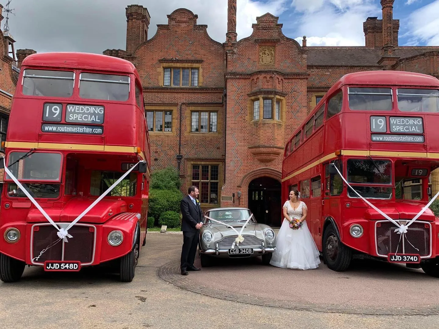 Two Matching Vintage Double Decker Red Routemaster Buses at a wedding venue with a Classic Aston Martin in the middle. Bride and Groom are standing by the buses posing for photos. All three vehicles have matching white ribbons